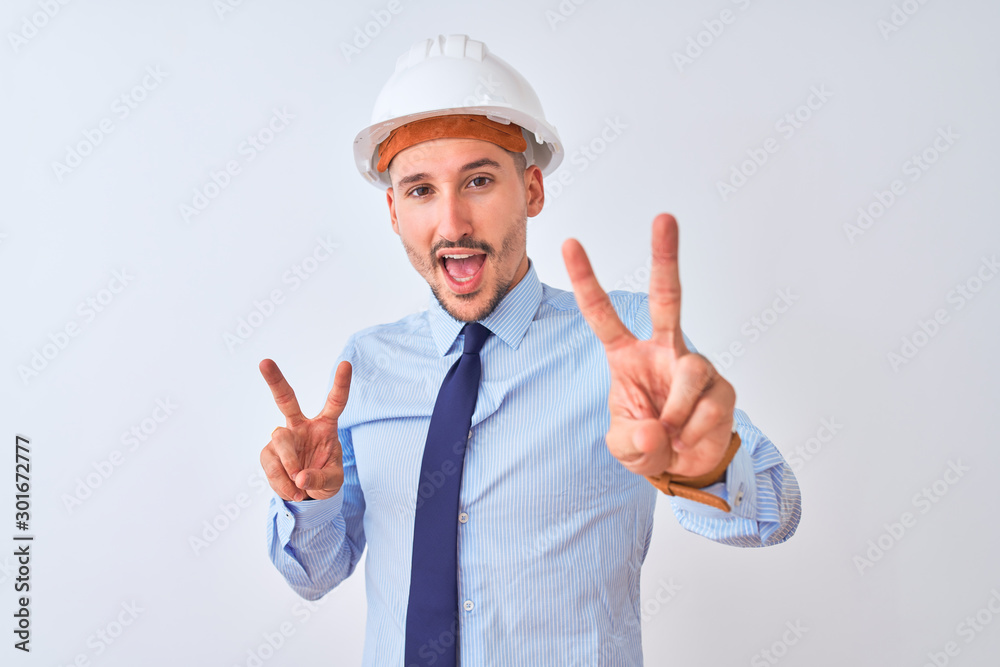 Young business man wearing contractor safety helmet over isolated background smiling looking to the camera showing fingers doing victory sign. Number two.