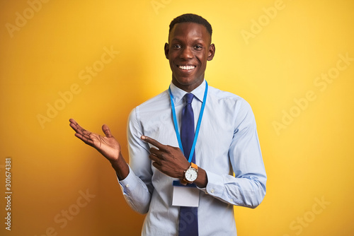 African american businessman wearing identification card over isolated yellow background amazed and smiling to the camera while presenting with hand and pointing with finger.