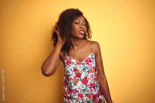African american woman wearing floral summer t-shirt over isolated yellow background smiling with hand over ear listening an hearing to rumor or gossip. Deafness concept.