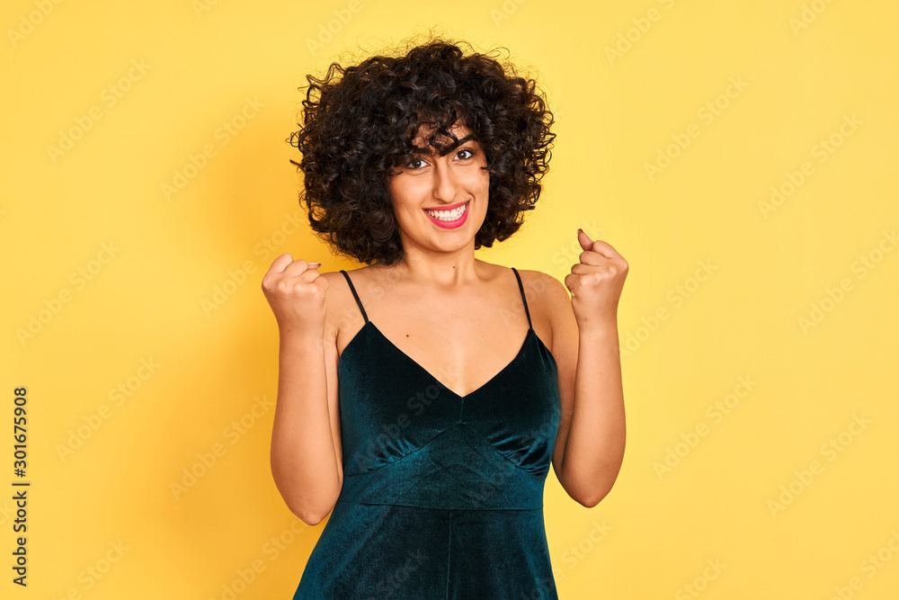Young arab woman with curly hair wearing elegant dress over isolated yellow background celebrating surprised and amazed for success with arms raised and open eyes. Winner concept. Young arab woman with curly hair wearing elegant dress over isolated yellow background celebrating surprised and amazed for success with arms raised and open eyes. Winner concept.