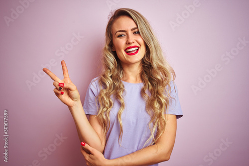 Young beautiful woman wearing purple t-shirt standing over pink isolated background smiling with happy face winking at the camera doing victory sign. Number two.