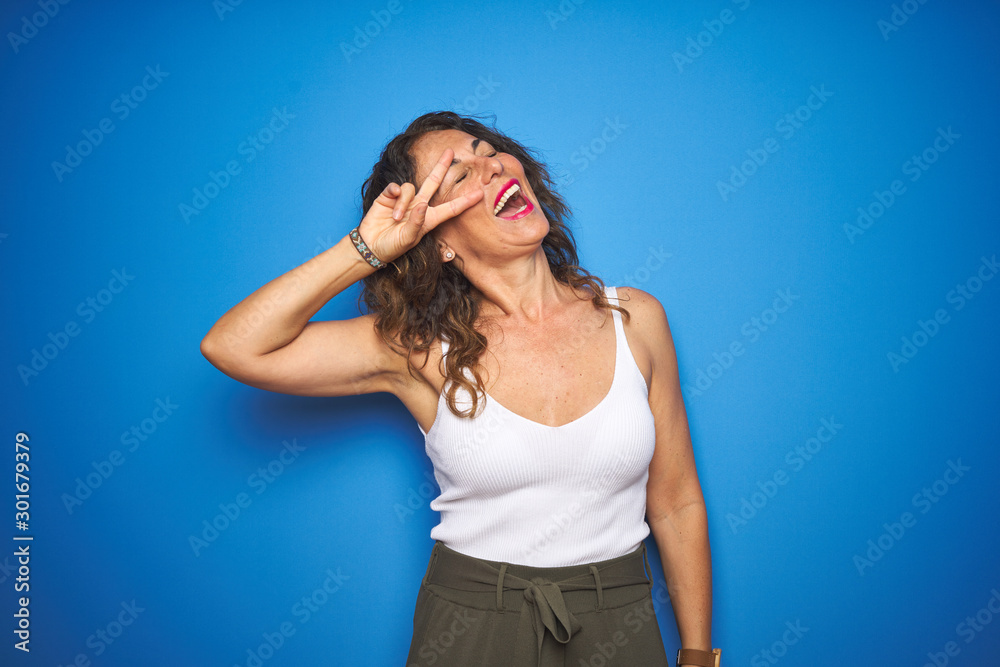 Middle age senior woman with curly hair standing over blue isolated background Doing peace symbol with fingers over face, smiling cheerful showing victory