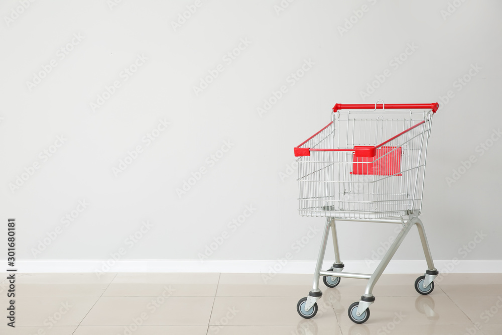 Empty shopping cart near light wall - obrazy, fototapety, plakaty 