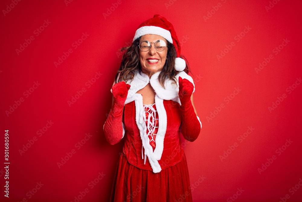 Middle age beautiful woman wearing Santa Claus costume over isolated red background excited for success with arms raised and eyes closed celebrating victory smiling. Winner concept. Middle age beautiful woman wearing Santa Claus costume over isolated red background excited for success with arms raised and eyes closed celebrating victory smiling. Winner concept.