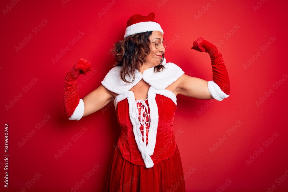 Middle age beautiful woman wearing Santa Claus costume over isolated red background showing arms muscles smiling proud. Fitness concept. Middle age beautiful woman wearing Santa Claus costume over isolated red background showing arms muscles smiling proud. Fitness concept.