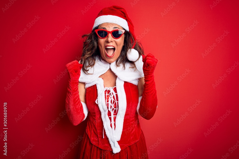 Middle age woman wearing Santa Claus costume and sunglasses over isolated red background celebrating surprised and amazed for success with arms raised and open eyes. Winner concept. Middle age woman wearing Santa Claus costume and sunglasses over isolated red background celebrating surprised and amazed for success with arms raised and open eyes. Winner concept.