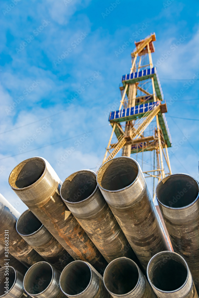 In the foreground pipes for drilling oil and gas wells. In the background, a blurred image of the rig out of focus. Blue sky with clouds. In the foreground pipes for drilling oil and gas wells. In the background, a blurred image of the rig out of focus. Blue sky with clouds.