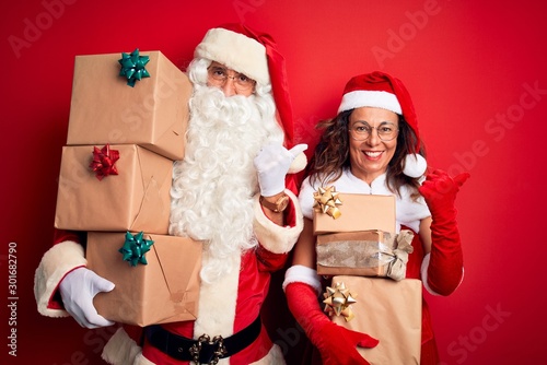 Middle age couple wearing Santa costume holding tower of gifts over isolated red background Pointing to the back behind with hand and thumbs up, smiling confident