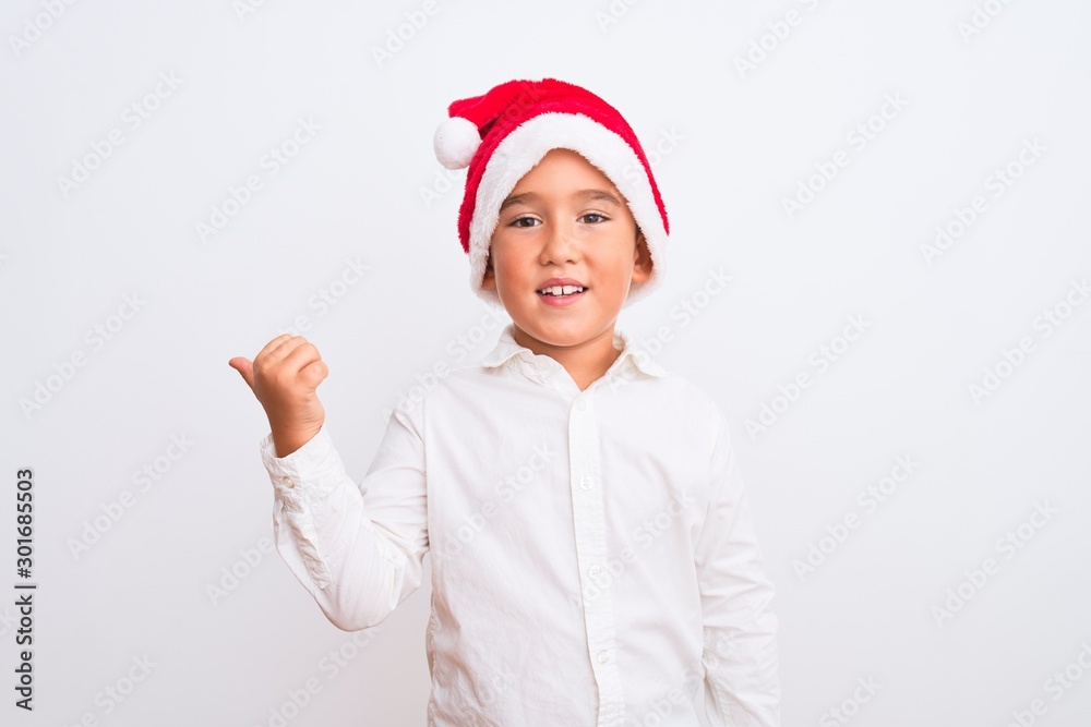 Beautiful kid boy wearing Christmas Santa hat standing over isolated white background smiling with happy face looking and pointing to the side with thumb up.