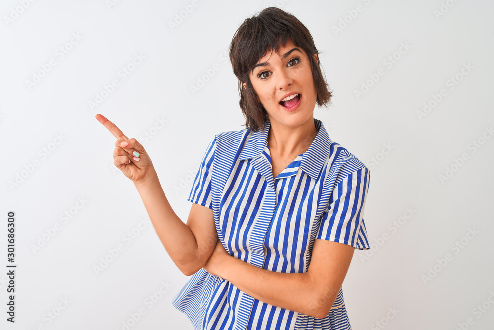 Young beautiful woman wearing blue striped shirt standing over isolated white background with a big smile on face, pointing with hand and finger to the side looking at the camera. Young beautiful woman wearing blue striped shirt standing over isolated white background with a big smile on face, pointing with hand and finger to the side looking at the camera.