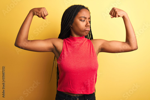 African american woman wearing red casual t-shirt standing over isolated yellow background showing arms muscles smiling proud. Fitness concept.