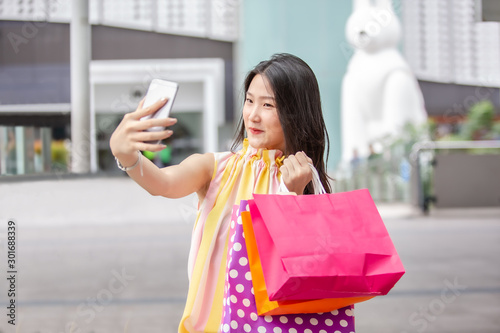 Young beautiful Asian woman holding her smartphone to taking a selfie with her shopping bags 