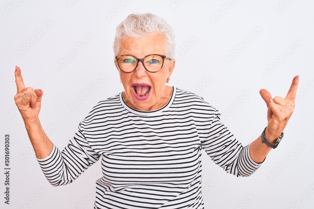 Senior grey-haired woman wearing striped navy t-shirt glasses over isolated white background shouting with crazy expression doing rock symbol with hands up. Music star. Heavy concept. Senior grey-haired woman wearing striped navy t-shirt glasses over isolated white background shouting with crazy expression doing rock symbol with hands up. Music star. Heavy concept.
