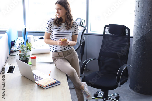 Attractive young woman holding smartphone and looking away while sitting on the desk in office.