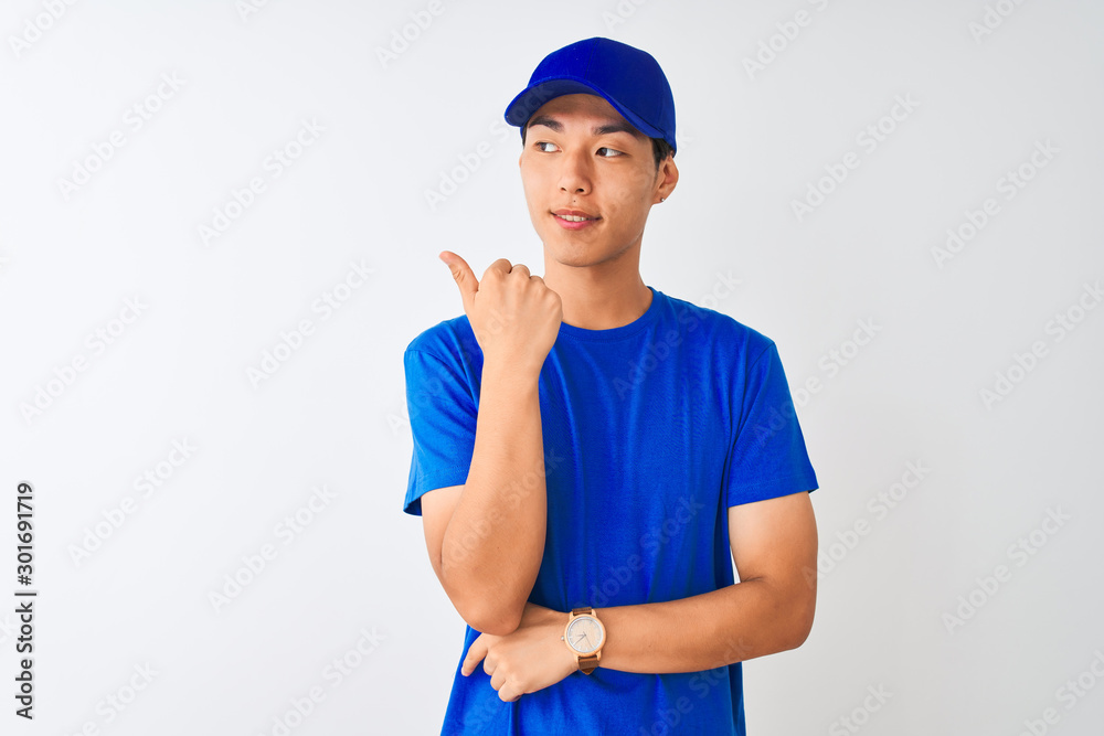 Chinese deliveryman wearing blue t-shirt and cap standing over isolated white background smiling with happy face looking and pointing to the side with thumb up.