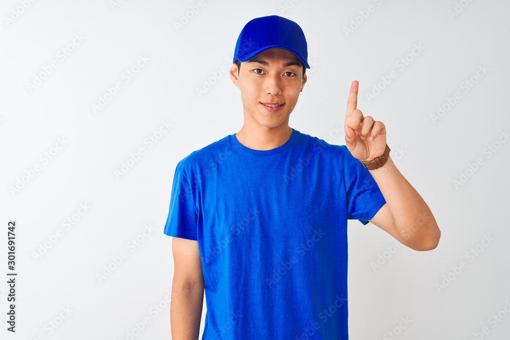 Chinese deliveryman wearing blue t-shirt and cap standing over isolated white background showing and pointing up with finger number one while smiling confident and happy.