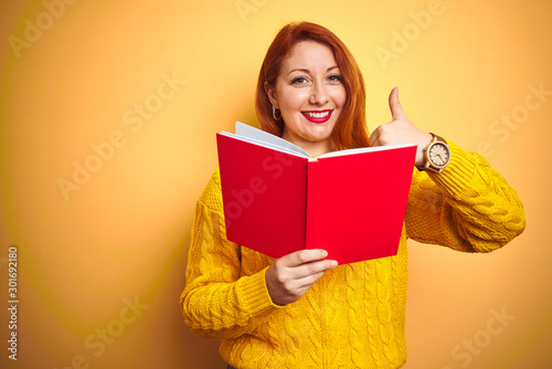 Young redhead teacher woman reading red book over yellow isolated background happy with big smile doing ok sign, thumb up with fingers, excellent sign