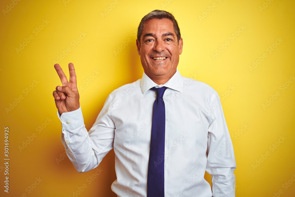 Handsome middle age businessman standing over isolated yellow background smiling with happy face winking at the camera doing victory sign. Number two. Handsome middle age businessman standing over isolated yellow background smiling with happy face winking at the camera doing victory sign. Number two.