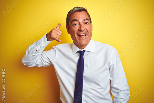 Handsome middle age businessman standing over isolated yellow background smiling doing phone gesture with hand and fingers like talking on the telephone. Communicating concepts.