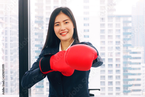 Young and cute Asian woman wearing suit and red boxing gloves in the office smiling and ready to fight with hard work and strong competition at work and in business concept © TS.PHOTOS