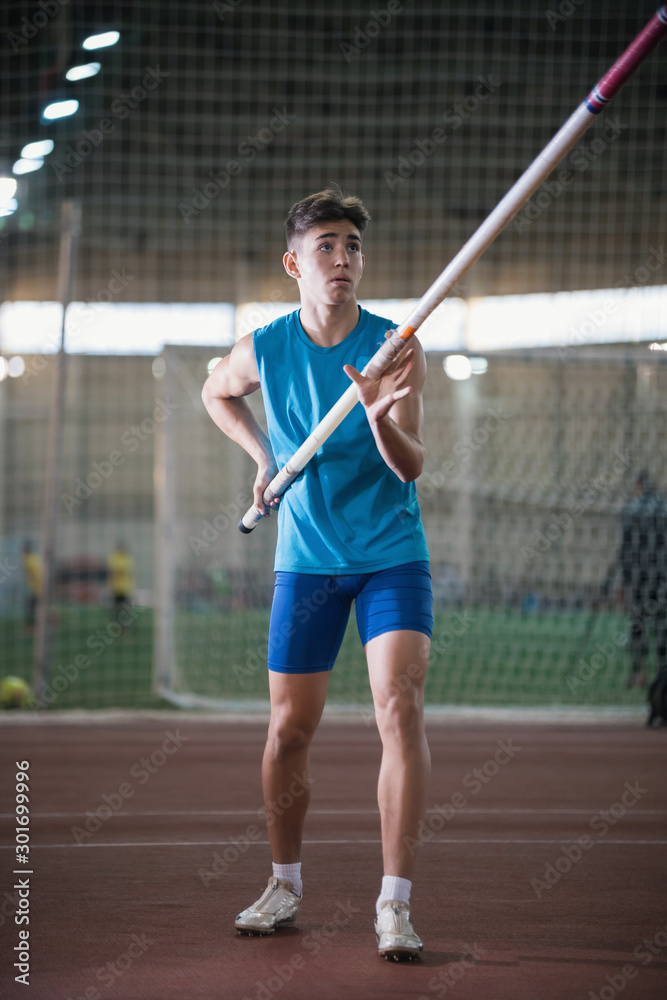 Pole vault training in the sports stadium - young man standing on the track holding a pole and looking up