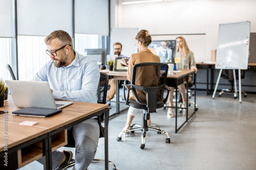 Senior man dressed casually working on the laptop in the office with coworkers on the background