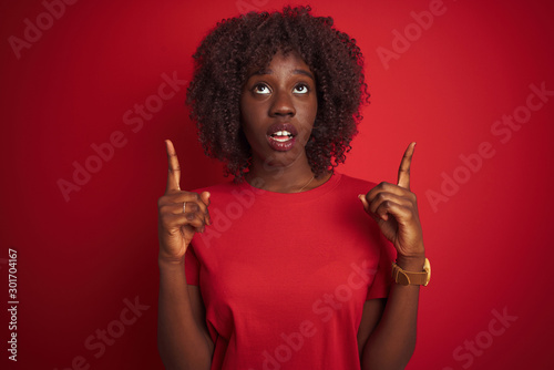 Young african afro woman wearing t-shirt standing over isolated red background amazed and surprised looking up and pointing with fingers and raised arms.