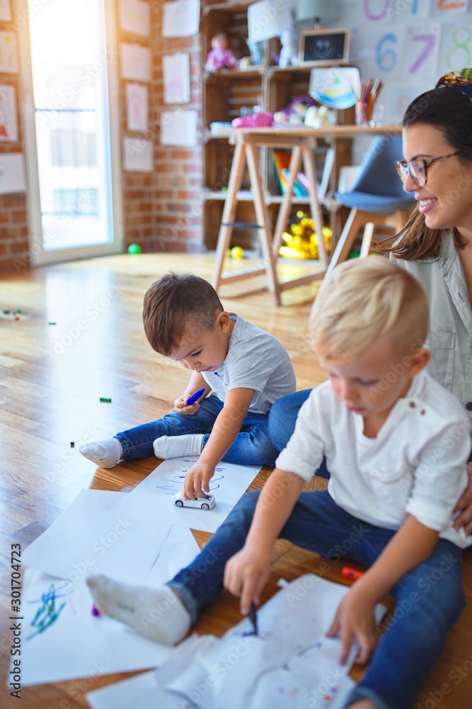 Obraz premium Beautiful teacher and group of toddlers sitting on the floor drawing using paper and pencil around lots of toys at kindergarten