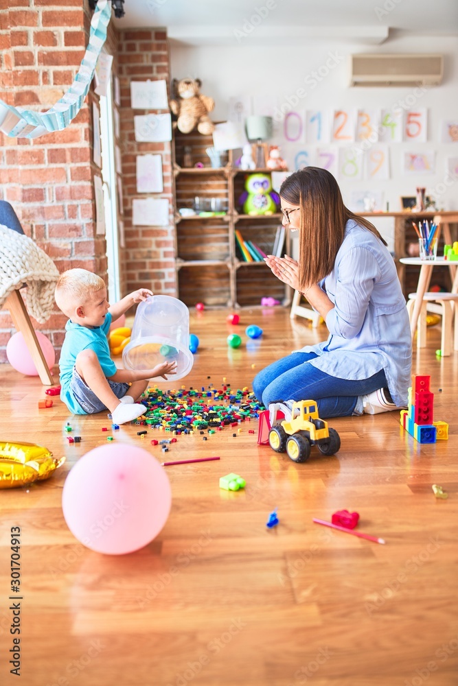 Fototapeta premium Young caucasian child playing at playschool with teacher. Mother and son at playroom bulding a tower with toy blocks