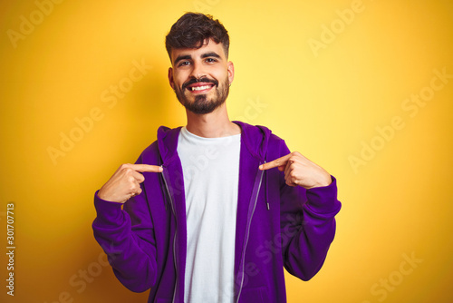 Young man with tattoo wearing sport purple sweatshirt over isolated yellow background looking confident with smile on face, pointing oneself with fingers proud and happy.
