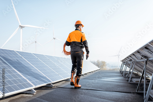 Well-equipped worker in protective orange clothing walking and examining solar panels on a photovoltaic rooftop plant. Concept of maintenance and installation of solar stations