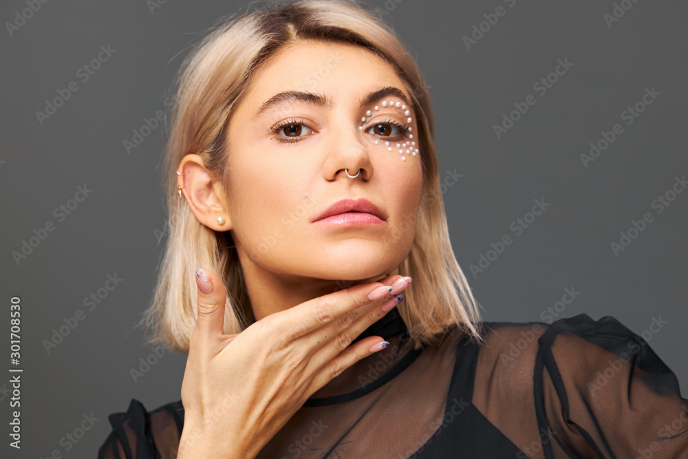 Pretty fashionable blonde woman with trendy glamourous make up posing isolated holding hand under chin, showing neat polished nails, looking at camera with confident enigmatic facial expression Pretty fashionable blonde woman with trendy glamourous make up posing isolated holding hand under chin, showing neat polished nails, looking at camera with confident enigmatic facial expression