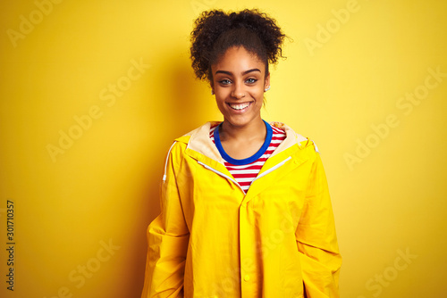Young african american woman wearing rain coat over isolated yellow background with a happy and cool smile on face. Lucky person.