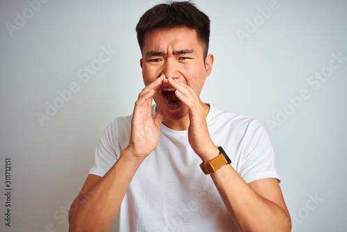 Young asian chinese man wearing t-shirt standing over isolated white background Shouting angry out loud with hands over mouth
