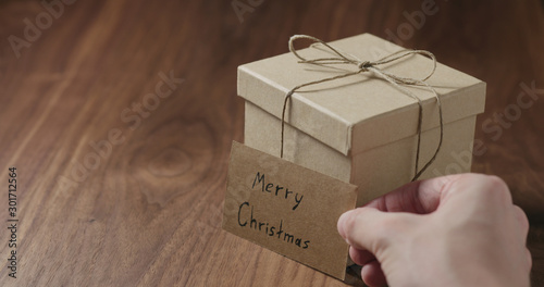 man hand put merry christmas card next to brown paper gift box on walnut table