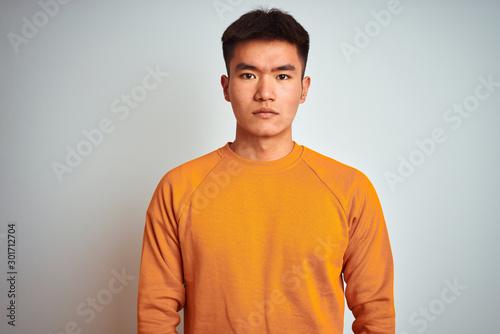 Young asian chinese man wearing orange sweater standing over isolated white background with serious expression on face. Simple and natural looking at the camera.