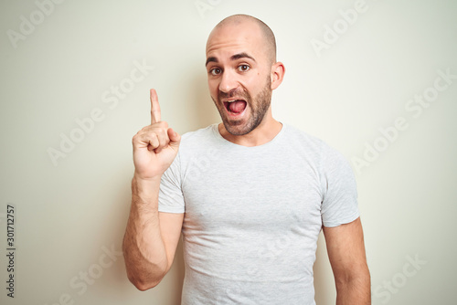 Young bald man with beard wearing casual white t-shirt over isolated background pointing finger up with successful idea. Exited and happy. Number one.