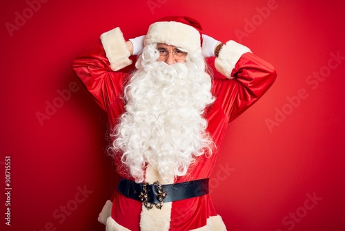 Middle age handsome man wearing Santa costume standing over isolated red background relaxing and stretching, arms and hands behind head and neck smiling happy