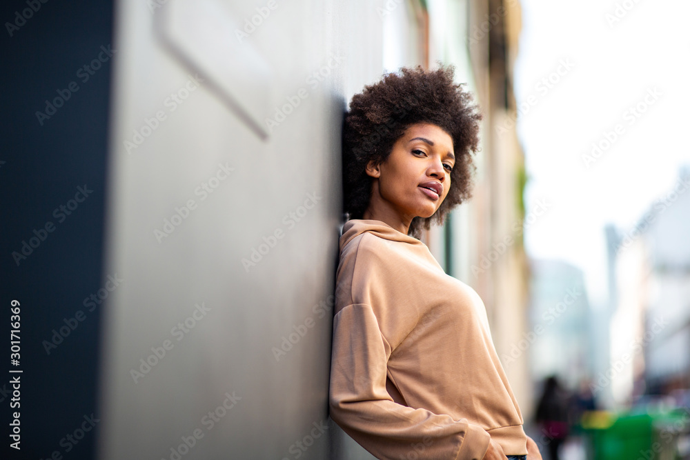 beautiful black female fashion model with afro hairstyle leaning against wall - obrazy, fototapety, plakaty 