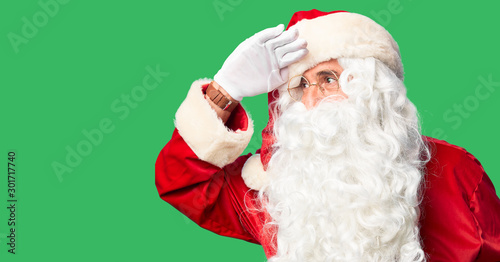Middle age handsome man wearing Santa Claus costume and beard standing very happy and smiling looking far away with hand over head. Searching concept.