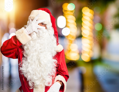 Middle age handsome man wearing Santa Claus costume and beard standing peeking in shock covering face and eyes with hand, looking through fingers with embarrassed expression.