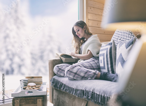 Young beautiful blonde woman with book sitting home in living room by the window. Winter snow landscape view.