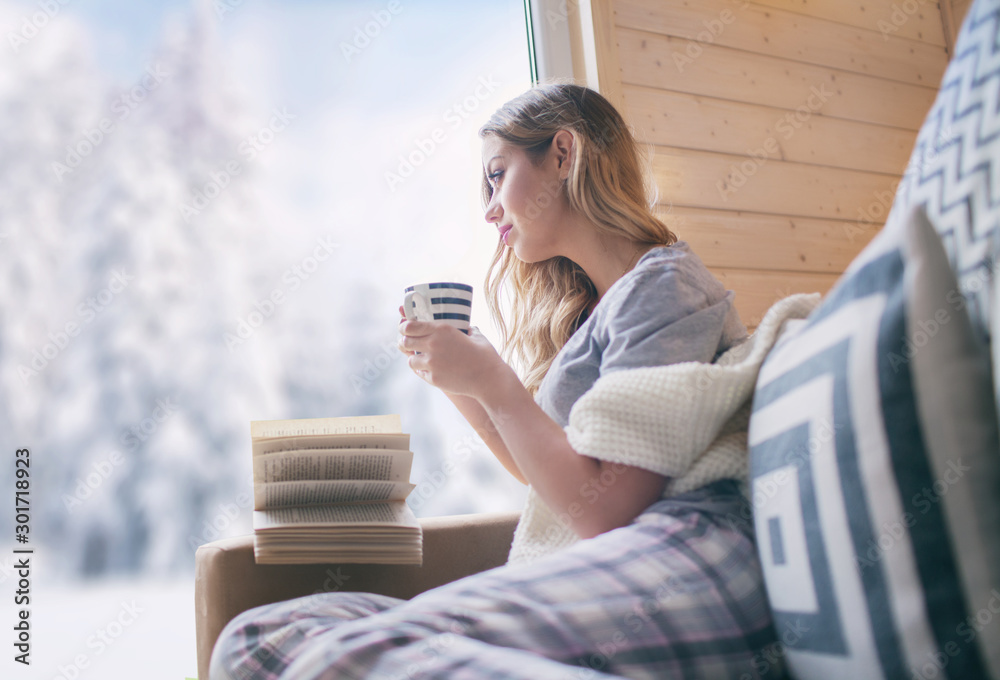 Young beautiful blonde woman with cup of coffee sitting home in living room by the window. Winter snow landscape view. Christmas concept  - obrazy, fototapety, plakaty 