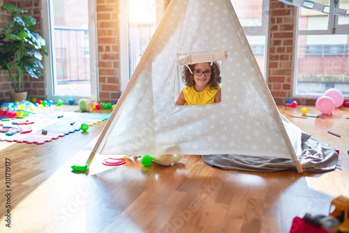 Beautiful toddler wearing glasses and unicorn diadem sitting on the floor inside tipi at kindergarten
