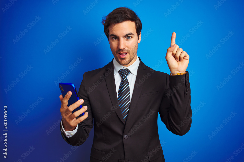 Young handsome businessman using smartphone standing over isolated blue background surprised with an idea or question pointing finger with happy face, number one Young handsome businessman using smartphone standing over isolated blue background surprised with an idea or question pointing finger with happy face, number one