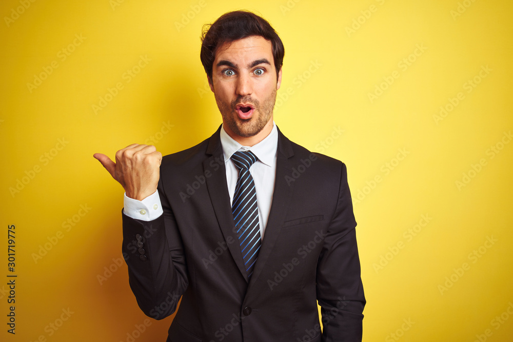 Young handsome businessman wearing suit and tie standing over isolated yellow background Surprised pointing with hand finger to the side, open mouth amazed expression. Young handsome businessman wearing suit and tie standing over isolated yellow background Surprised pointing with hand finger to the side, open mouth amazed expression.