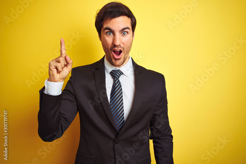 Young handsome businessman wearing suit and tie standing over isolated yellow background pointing finger up with successful idea. Exited and happy. Number one.