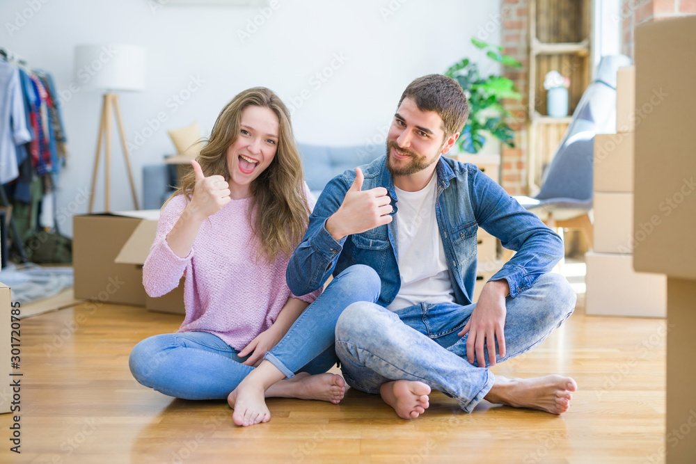 Young beautiful couple moving to a new house sitting on the floor doing happy thumbs up gesture with hand. Approving expression looking at the camera with showing success. Young beautiful couple moving to a new house sitting on the floor doing happy thumbs up gesture with hand. Approving expression looking at the camera with showing success.