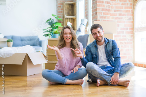 Young beautiful couple moving to a new house sitting on the floor amazed and smiling to the camera while presenting with hand and pointing with finger.