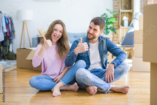 Young beautiful couple moving to a new house sitting on the floor doing happy thumbs up gesture with hand. Approving expression looking at the camera with showing success.